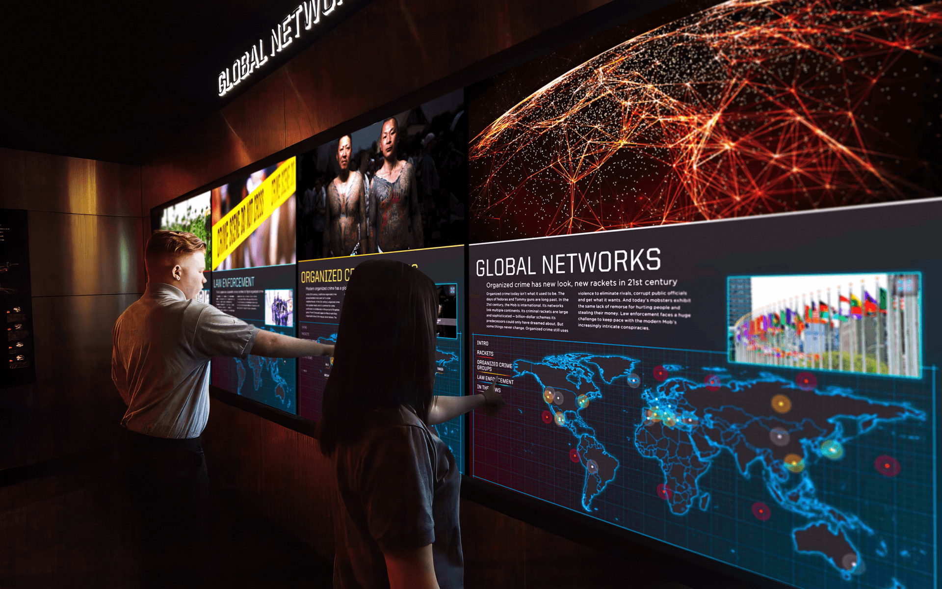Two museum visitors stand in front of a video wall displaying information about global organized crime. Each visitor is interacting with the exhibit by touching the screen.