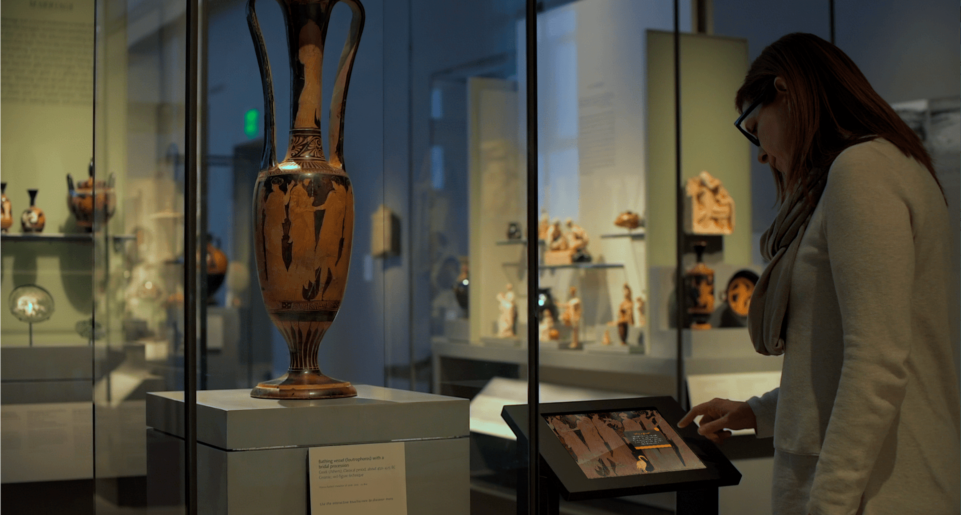 A museum visitor interacts with a touch screen next to a glass case containing a classical greek vessel. The screen shows a section of the red-figure imagery from the vessel. One figure on the screen is highlighted and accompanied by an info popup.